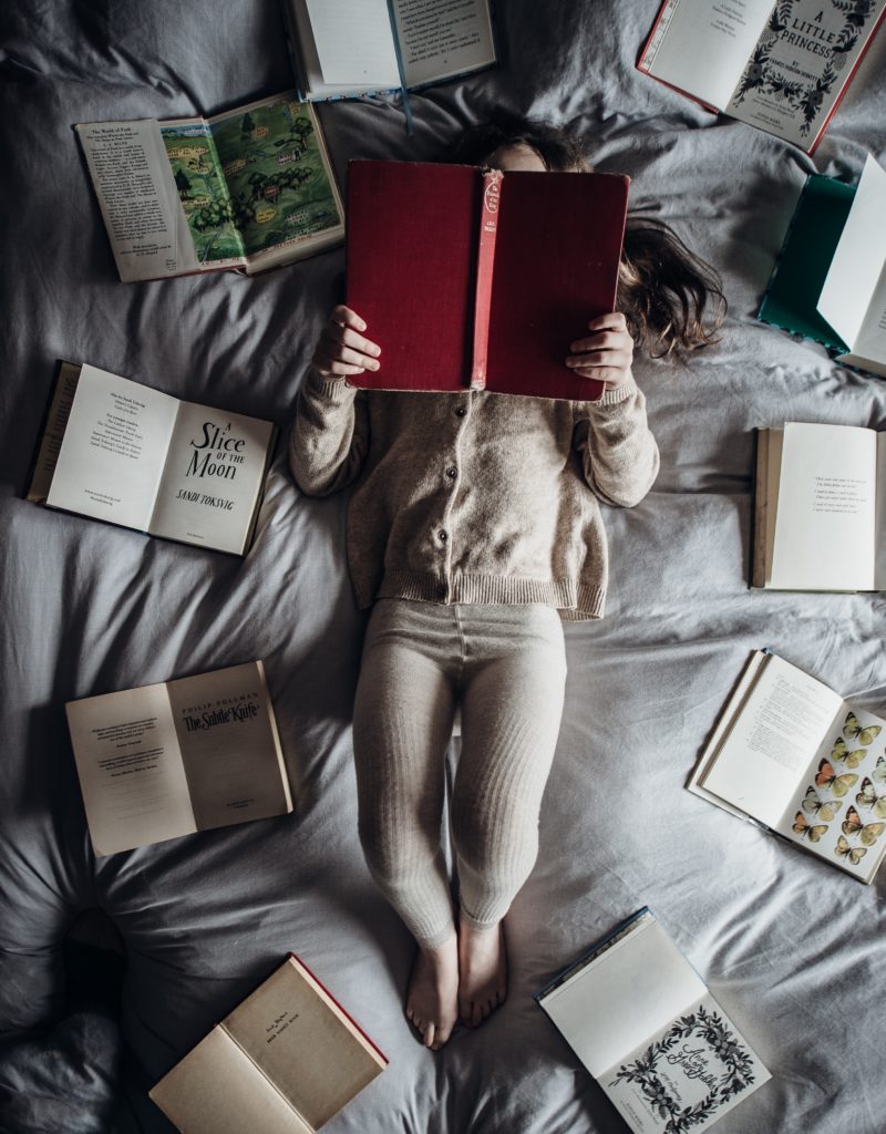child reading a book while surrounded by books on a bed