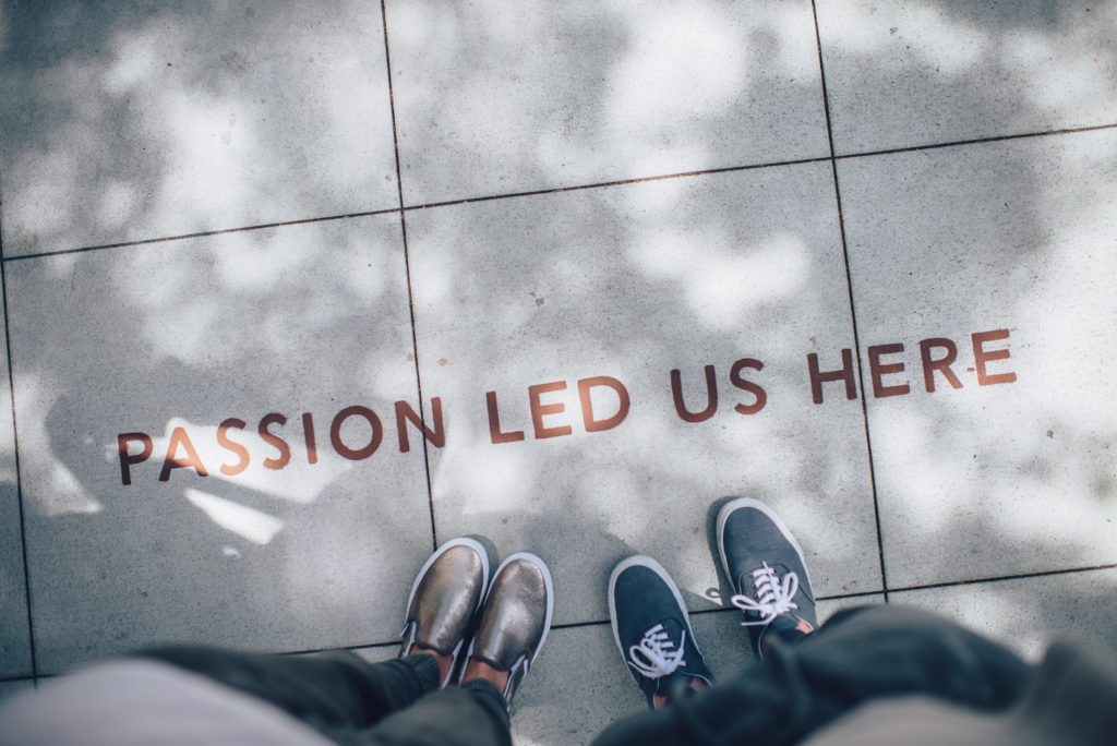 Image of a sidewalk with the words "Passion led us here" imprinted on it with the feet of two people visible standing below the sentence.